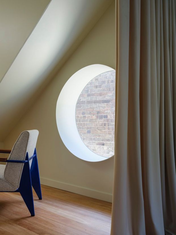 A softly lit interior corner featuring a distinctive circular window set into a pale wall, with a brick wall visible outside. The room has warm timber flooring, floor-to-ceiling curtains, and part of a sculptural blue and grey chair. A sloping ceiling and carefully framed natural light create a calm, contemporary atmosphere.
