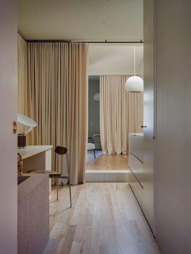 A refined interior view looking along a light timber-floored hallway or dressing area toward a raised room. Full-height cream curtains form soft partitions and are reflected in a large mirrored or glossy wardrobe surface on the right. A compact desk with a sculptural white lamp and dark chair sits to the left, while a spherical pendant light hangs in the background. The palette is warm and muted, with pale wood, cream textiles, soft beige cabinetry, and subtle pink-brown furnishings.