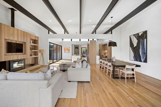 Bright open-plan contemporary interior combining a living room, dining area, and kitchen. The space features pale timber flooring, a large light-coloured sectional sofa, built-in timber media cabinetry with fireplace, exposed dark ceiling beams, a long timber dining table with woven chairs, and extensive glazing at the rear.