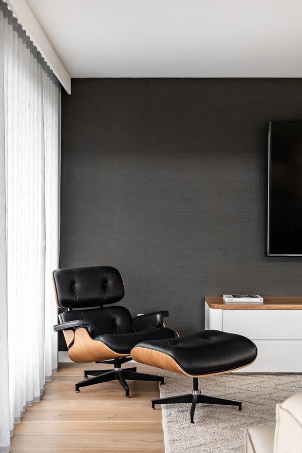 A refined living room corner featuring a black leather lounge chair and matching ottoman with warm timber shells, set against a dark textured feature wall. A wall-mounted television, low white and timber media console, pale timber flooring, sheer full-height curtains, and a neutral textured rug complete the contemporary interior.