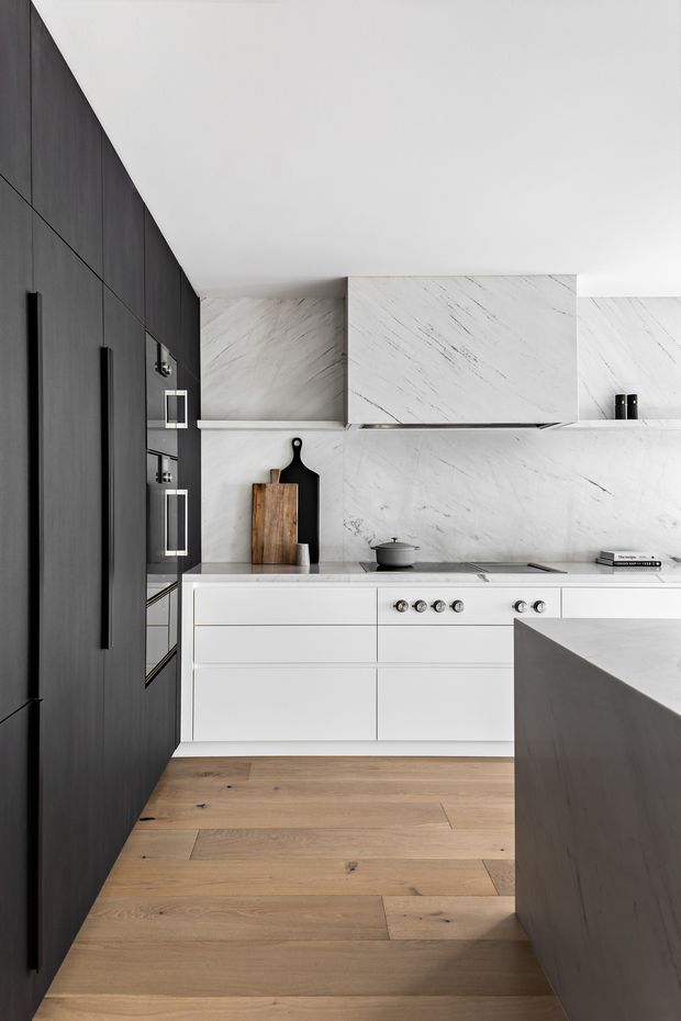 A refined contemporary kitchen with a white veined stone backsplash and matching upper cabinetry, white flat-panel base cabinets, integrated appliances, and a dark charcoal-toned full-height cabinet wall. Warm natural timber plank flooring contrasts with the monochromatic cabinetry. A dark island edge occupies the foreground, with simple timber and black cutting boards displayed on the benchtop.