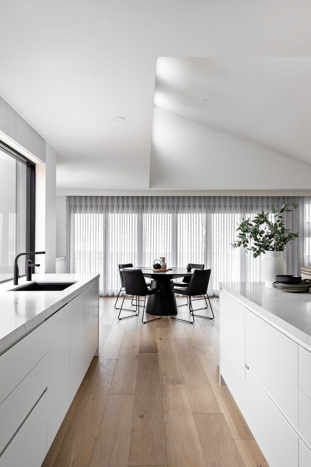 Bright, minimalist open-plan kitchen and dining area with white handleless cabinetry, a large island with black tap and sink, pale timber flooring, and a round black dining table with four black chairs. Full-height sheer curtains and large windows provide abundant diffuse natural light. A white benchtop and restrained black accents create a clean contemporary interior.