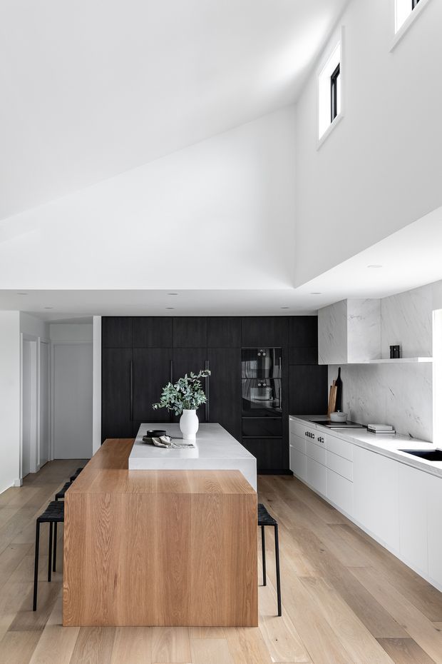 A bright, minimalist contemporary kitchen with a large timber-clad island and integrated seating. The room features pale timber flooring, matte black full-height cabinetry with integrated appliances, white cabinetry, and a pale veined stone splashback and benchtop. A dramatic angled ceiling void with high clerestory-style windows floods the space with natural light.