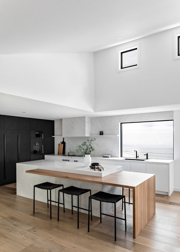 A bright contemporary kitchen with a large island featuring a white stone benchtop and timber waterfall-style side panels. Three black bar stools sit along the island. The kitchen includes dark timber cabinetry, white marble-look splashback and wall surfaces, integrated appliances, pale timber flooring, and a large window overlooking the ocean. A high ceiling with clerestory windows adds openness and natural light.