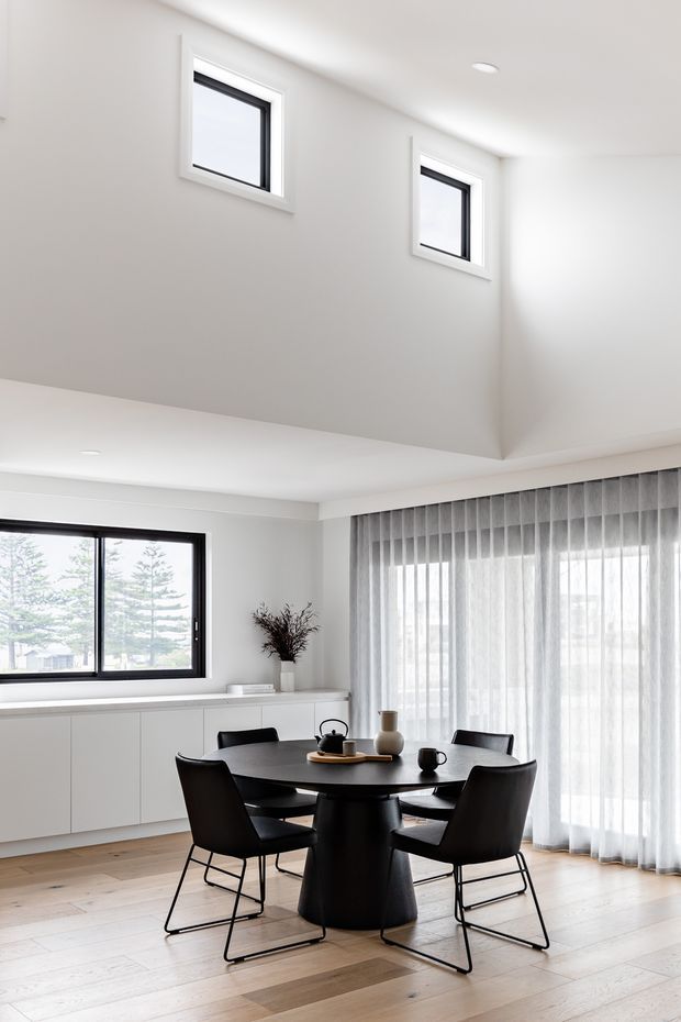 Bright contemporary dining area with a round black dining table and four black chairs, set against white built-in cabinetry and large windows with sheer curtains. Pale timber flooring, minimal decor, and a high ceiling with clerestory windows create an airy, light-filled interior.