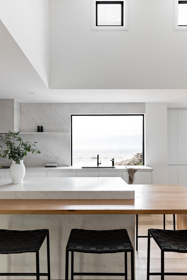 Bright, minimalist contemporary kitchen with a large island and integrated dining-height timber counter. White cabinetry, pale veined stone splashback and benchtops, black-framed glazing, and black woven bar stools create a restrained monochrome palette. A coastal view is visible through the main window, while high-level windows and a raised ceiling add openness.