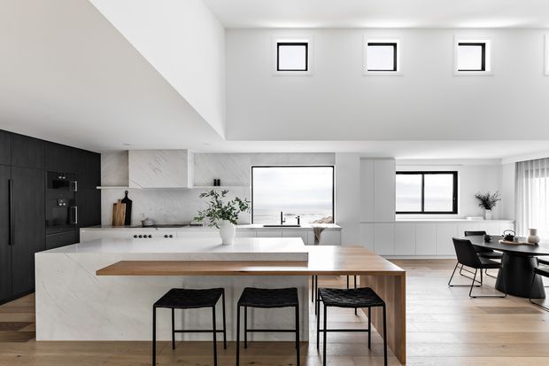 Bright contemporary open-plan kitchen and dining area with a large white marble-look island, integrated dark cabinetry, timber breakfast bar, black stools, pale timber flooring, and a dining table beside expansive windows. The room features a high sloping ceiling with a row of small clerestory windows and abundant natural light.
