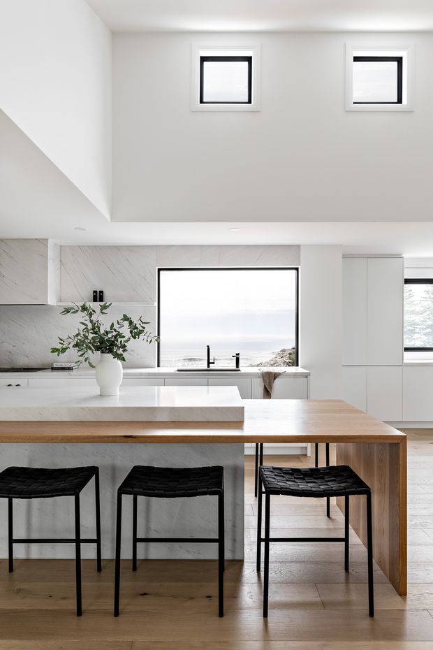 Bright contemporary kitchen with a large white stone island, integrated cabinetry, marble-look backsplash, timber dining extension, black metal-framed stools, and pale timber flooring. Large windows provide a coastal view, while a raised ceiling with clerestory-style square windows adds light and volume.