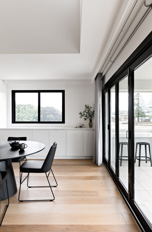 Bright, refined dining area with a round dark dining table and black upholstered chairs, set against white built-in cabinetry and large black-framed windows and sliding glass doors. Pale timber flooring, sheer curtains, a simple tea service, and a small potted plant create a minimalist contemporary interior with strong indoor–outdoor connection. A covered terrace with stools is visible beyond the glazing.