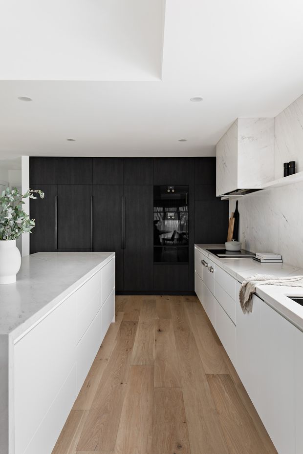 A refined contemporary kitchen viewed down a central aisle, with a large white island and waterfall-style benchtop in the foreground, white perimeter cabinetry, a dramatic dark timber or black pantry wall at the rear, and a veined white stone splashback and overhead cabinetry. Light timber flooring, recessed ceiling lights, integrated appliances, and restrained styling create a clean, high-contrast monochromatic scheme.