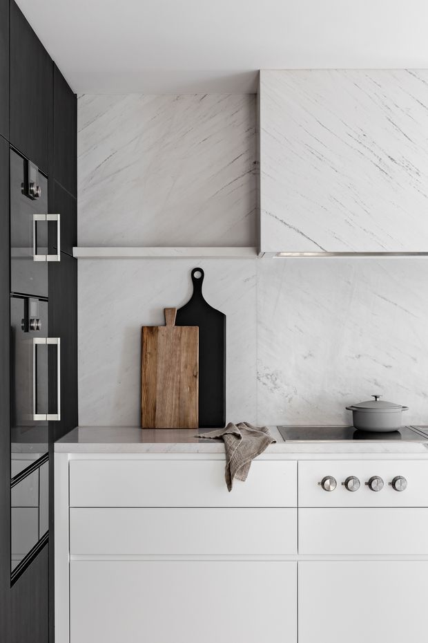 A close-up view of a refined contemporary kitchen with white marble-look slab cabinetry and backsplash, integrated dark cabinetry at the side, a pale stone benchtop, and a built-in cooktop and oven. Minimal black and timber cutting boards lean against the splashback, while a neutral linen cloth and grey lidded pot add understated texture.