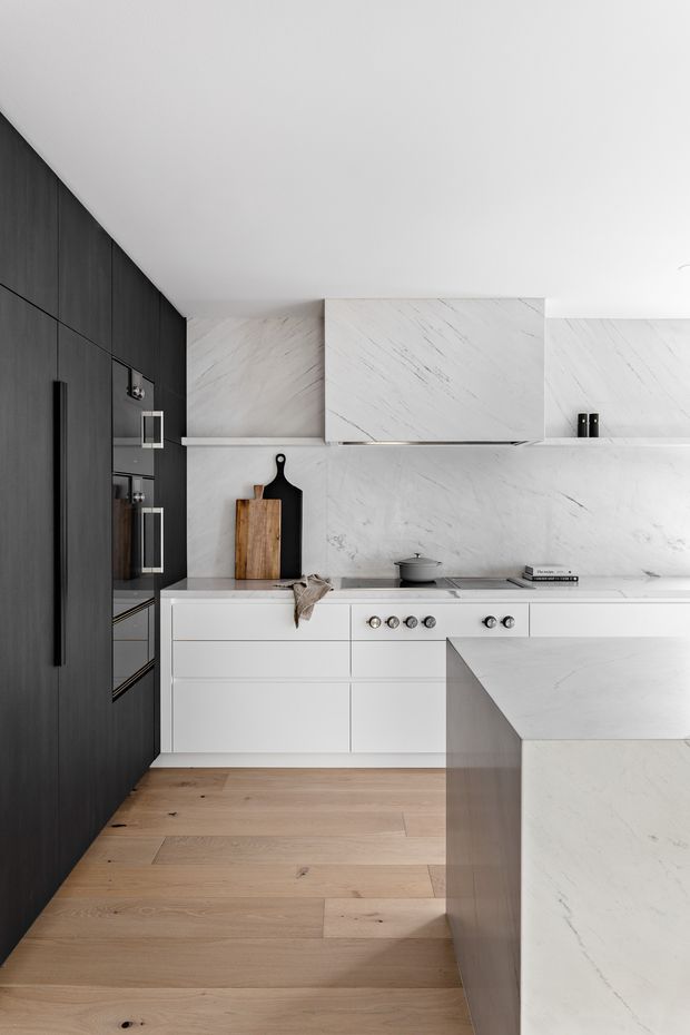 A refined contemporary kitchen with a minimalist black-and-white palette. The space features dark full-height cabinetry with integrated appliances, white marble-look cabinetry and splashback, a matching island, slim stainless-steel cooktop and oven controls, and wide light timber flooring. The clean-lined composition is softly lit and uncluttered.