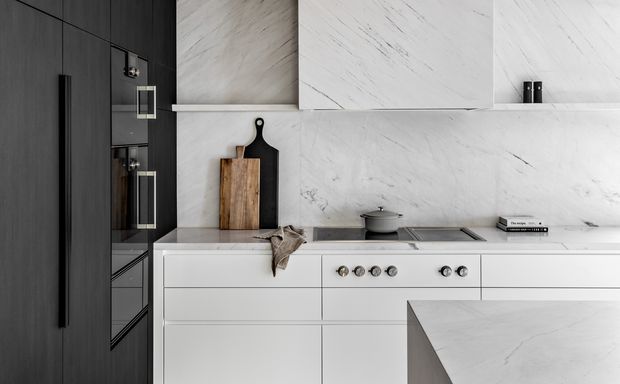 A refined contemporary kitchen featuring a long white stone benchtop and matching marble-look splashback, integrated cabinetry, a dark charcoal full-height cabinet bank, and a built-in cooktop and oven. Minimal black hardware, timber chopping boards, and a small grey pot add restrained contrast. A projecting island or peninsula occupies the foreground.