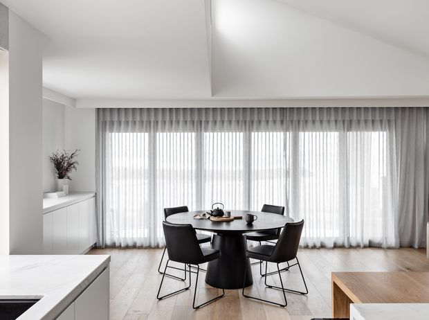 Bright contemporary dining area with a round dark dining table and matching black chairs positioned in front of full-height sheer curtains. Pale timber flooring, white walls and ceiling, streamlined white cabinetry, and a light stone kitchen island are visible, creating a clean open-plan interior.
