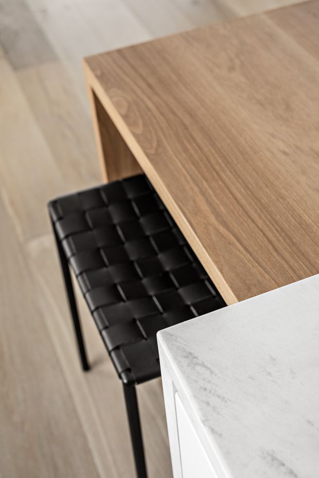 Close-up interior detail showing a warm timber dining or kitchen table with a black woven leather-style chair beside it. A white marble-look or stone benchtop occupies the foreground, and pale timber flooring runs throughout. The image highlights refined, contemporary material combinations and furniture detailing.