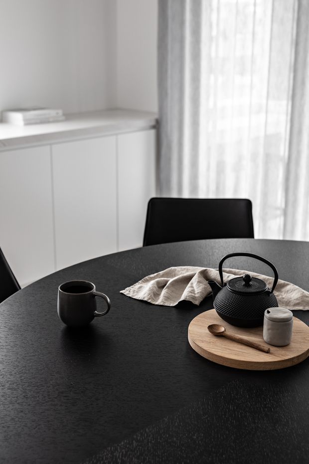 A softly lit minimalist dining area with a round black dining table, black upholstered chairs, a black ceramic mug, and a textured black cast-iron teapot arranged on a round timber serving board. White built-in cabinetry and sheer curtains create a calm, monochromatic interior.