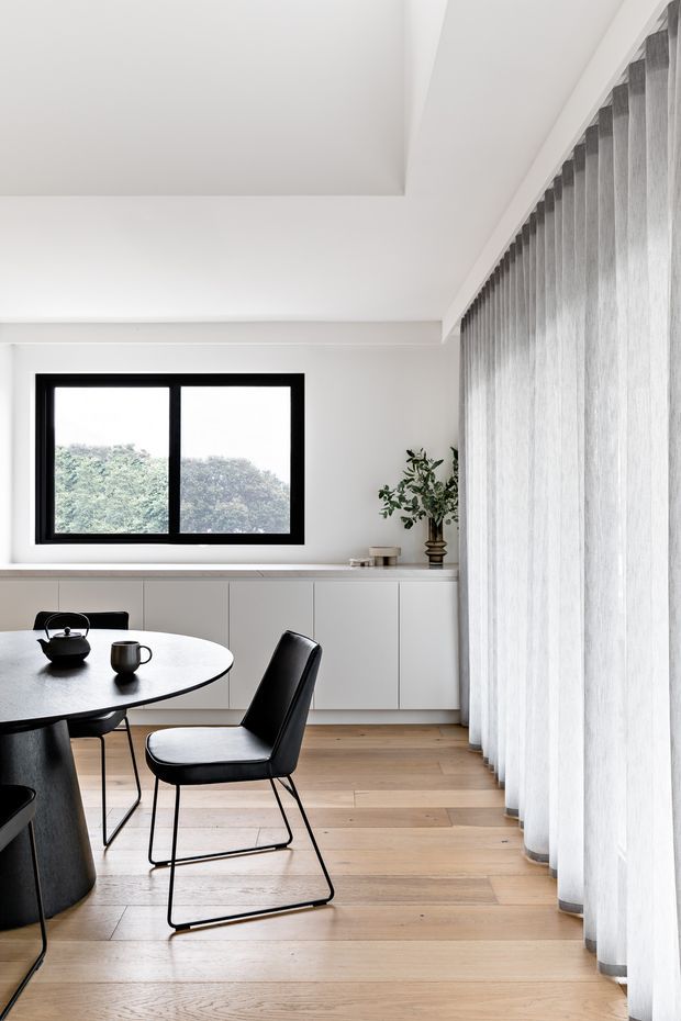 A bright, minimalist dining area with a round light-toned table, black dining chairs, white built-in cabinetry, black-framed window, sheer floor-to-ceiling curtains, and pale timber flooring. A small plant and simple black tableware add restrained accents, while a leafy outdoor view is visible through the window.