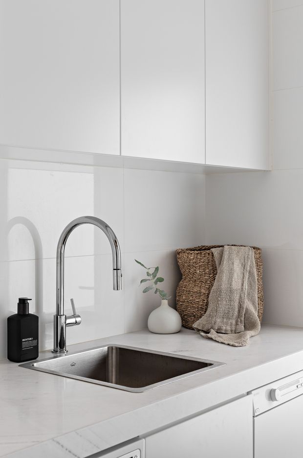 Bright, minimalist white kitchen sink area with seamless white upper cabinetry, white tiled splashback, pale stone benchtop, stainless-steel undermount sink and tall chrome gooseneck tap. A black soap dispenser, woven basket, neutral linen towel and small vase provide restrained accents.
