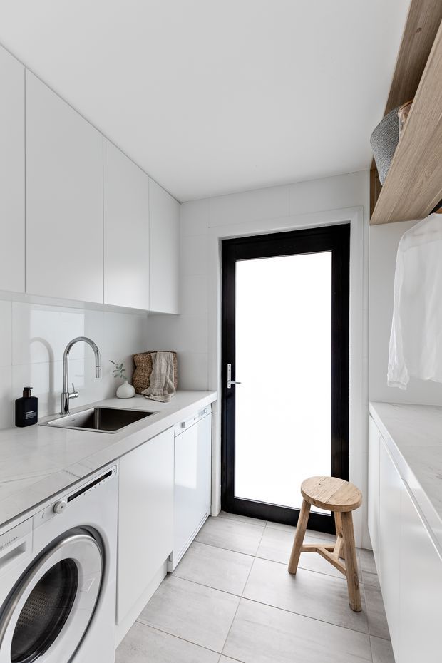 Bright, compact contemporary laundry room with white floor-to-ceiling cabinetry, a white stone-look benchtop, integrated stainless-steel sink and tapware, front-loading washing machine, and a black-framed glazed door. A timber open wardrobe unit with hanging clothes is visible on the right, along with a simple wooden stool. Large pale floor tiles and white surfaces create a clean, minimalist feel.