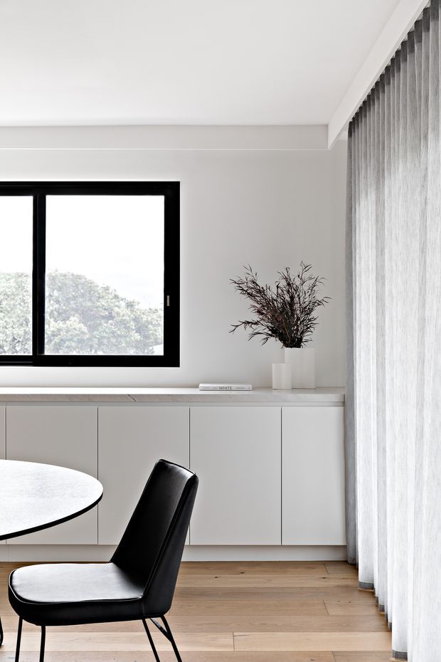 A bright, minimalist dining area with a round white table and black upholstered dining chairs beside a wall of sheer curtains. The room features pale timber flooring, white built-in cabinetry with a light stone or composite benchtop, a black-framed window, and simple white walls. A restrained vase arrangement provides a natural decorative accent.