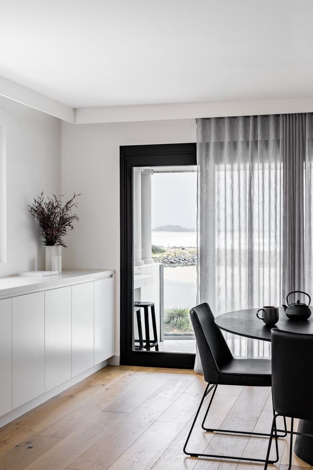 A refined dining area with a round dark dining table and black upholstered chairs beside a full-height glazed door. White built-in cabinetry lines the wall, while sheer grey curtains frame views toward a rocky coastal landscape. The room features pale timber flooring, white walls and ceiling, and restrained black accents in the joinery, door frame, furniture and tableware.