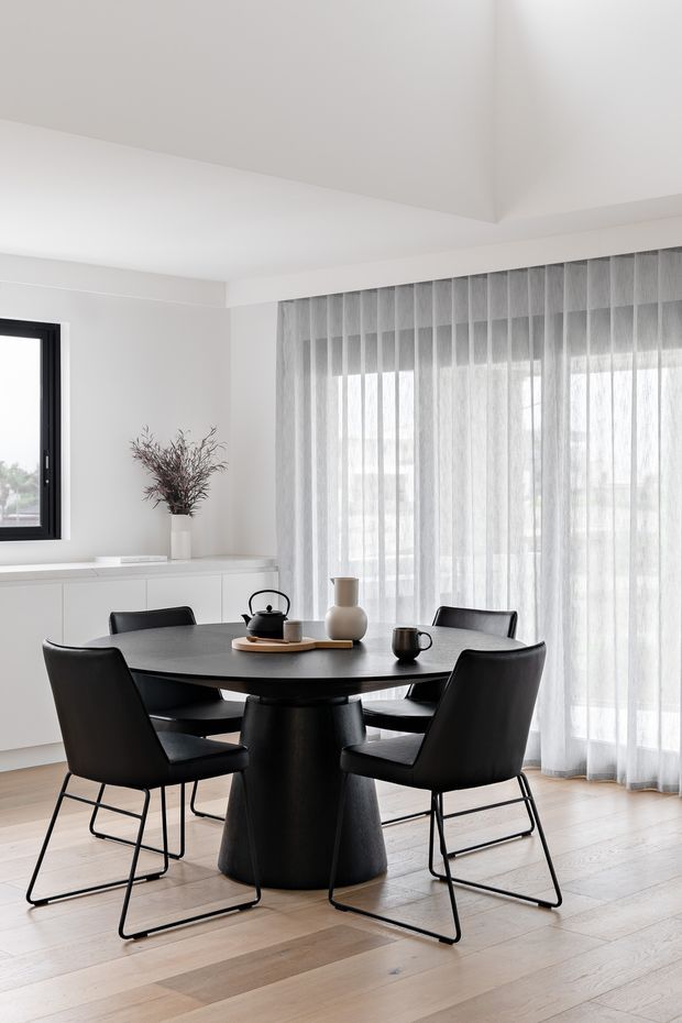 Bright, minimalist dining area with a round black dining table and six black upholstered chairs on light timber flooring. Sheer floor-to-ceiling curtains cover large glazing, while white built-in cabinetry and a simple vase arrangement line the rear wall. The space has a clean, contemporary monochrome palette and abundant natural light.