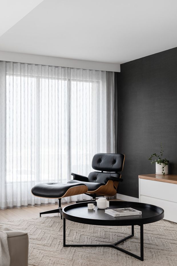 A refined contemporary living room with a black leather lounge chair and matching ottoman positioned beside a large window dressed in sheer white curtains. The room features a dark textured feature wall, pale built-in cabinetry with a timber-toned top, a round black coffee table, and a light textured area rug. The palette is monochromatic with warm timber accents and soft natural light.