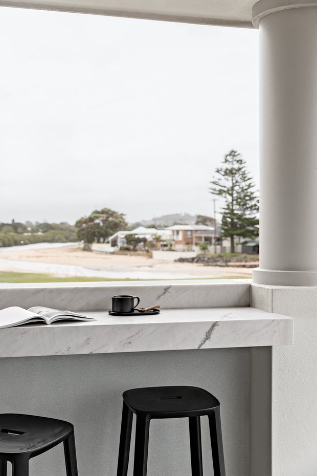 A minimalist coastal breakfast bar or outdoor entertaining counter beneath a covered terrace. The pale stone counter and backsplash frame a beach and residential view, with two black stools, a coffee cup and an open magazine. A large rounded column and simple plastered surfaces create a clean, refined architectural setting.