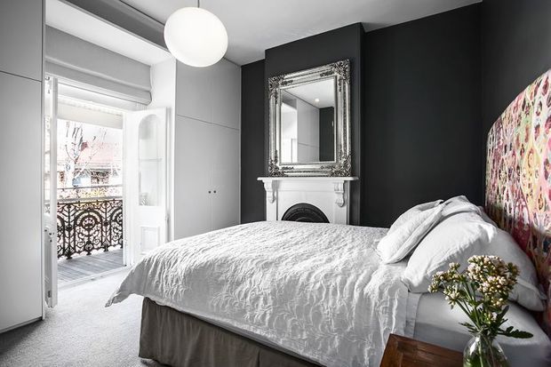 A refined bedroom with a white upholstered or timber bed dressed in textured white bedding, set against a dramatic black feature wall. The wall includes an ornate white fireplace surround and large decorative framed mirror. Floor-to-ceiling grey cabinetry occupies one side, while glazed French doors open to a balcony with decorative metal railing. A colourful patterned artwork or upholstered feature panel adds contrast at the head of the bed, complemented by a simple spherical pendant light and fresh flowers.
