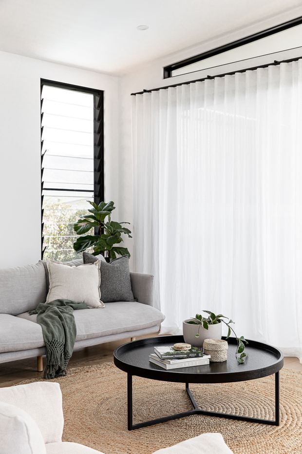 Bright, serene living room with a light grey sofa, textured neutral rug, black round coffee table, soft white sheer curtains, black-framed windows, and indoor plants. The room has a clean contemporary feel with abundant natural light and a restrained neutral palette.