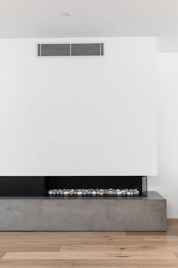 Minimalist interior detail featuring a broad white wall with a discreet linear air vent above a recessed black fireplace niche. The fireplace is set within a substantial smooth grey concrete surround, with decorative pale stones inside, and sits above a light natural timber floor. The composition is clean, restrained, and contemporary.