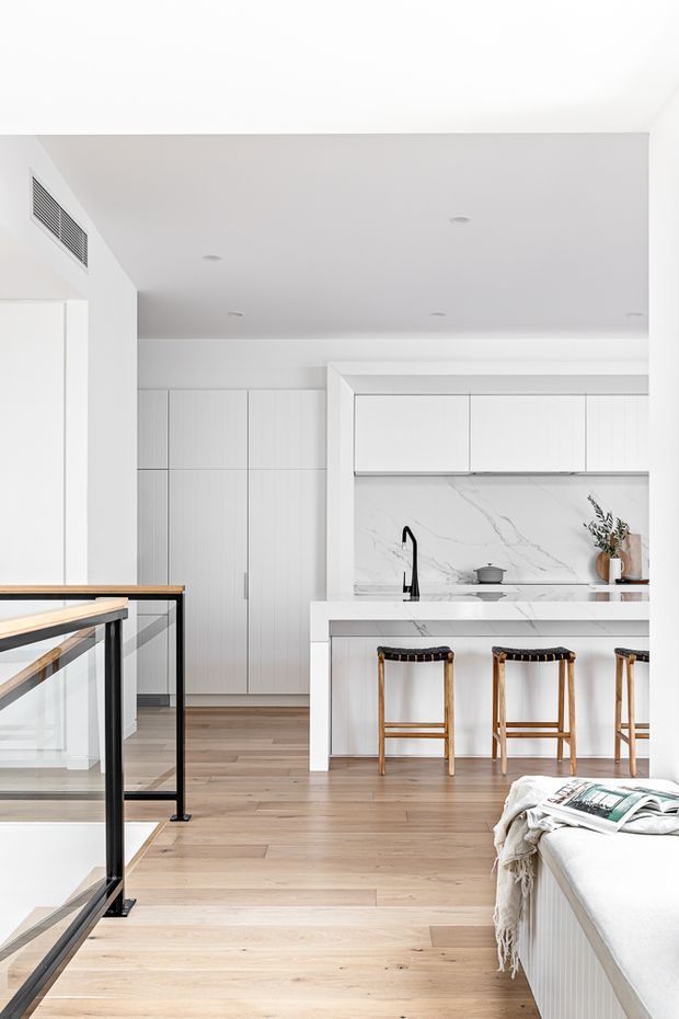 Bright open-plan kitchen and living area with white floor-to-ceiling cabinetry, a marble-look splashback, white island bench with integrated sink and black tapware, and timber bar stools. Pale timber flooring, white walls and ceiling, and a black-framed stair balustrade create a clean contemporary interior. A partial sofa or upholstered bench is visible in the foreground.