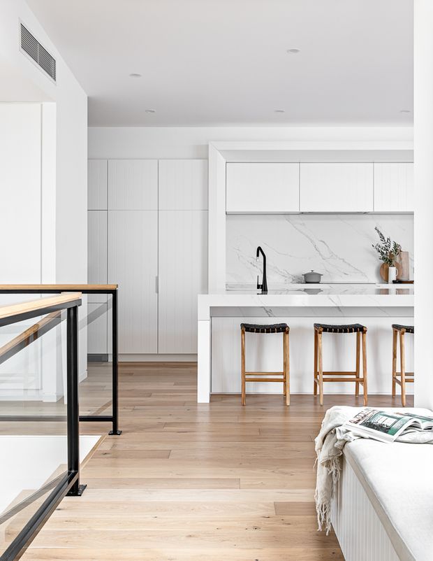 Bright, minimalist open-plan kitchen viewed from an adjacent living area, with white full-height cabinetry, a marble-look splashback, white island with black tap, timber bar stools, light timber flooring, and black-framed glass stair balustrade. A partial sofa and coffee-table reading material are visible in the foreground.