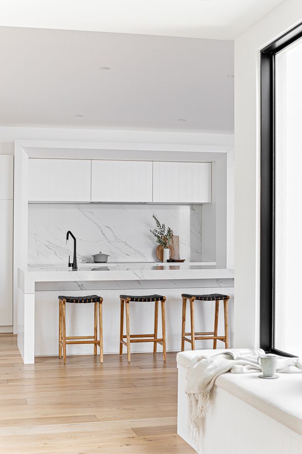 Bright, minimalist open-plan kitchen with a white island and integrated breakfast bar, three woven-seat timber stools, white vertically panelled cabinetry, and a dramatic white marble-look splashback with grey veining. Pale timber flooring, recessed ceiling lights, black-framed full-height glazing, and a built-in window seat create an airy, refined interior.