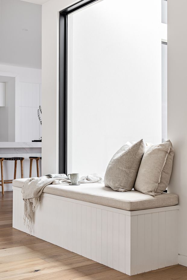 Bright, minimalist interior featuring a built-in window seat with white vertical panelling, a thick upholstered cushion, neutral textured pillows, and a draped throw. A large black-framed window provides strong natural light, while part of an adjacent kitchen with a stone benchtop and timber stools is visible in the background. Light timber flooring and soft white finishes create an airy, calm atmosphere.