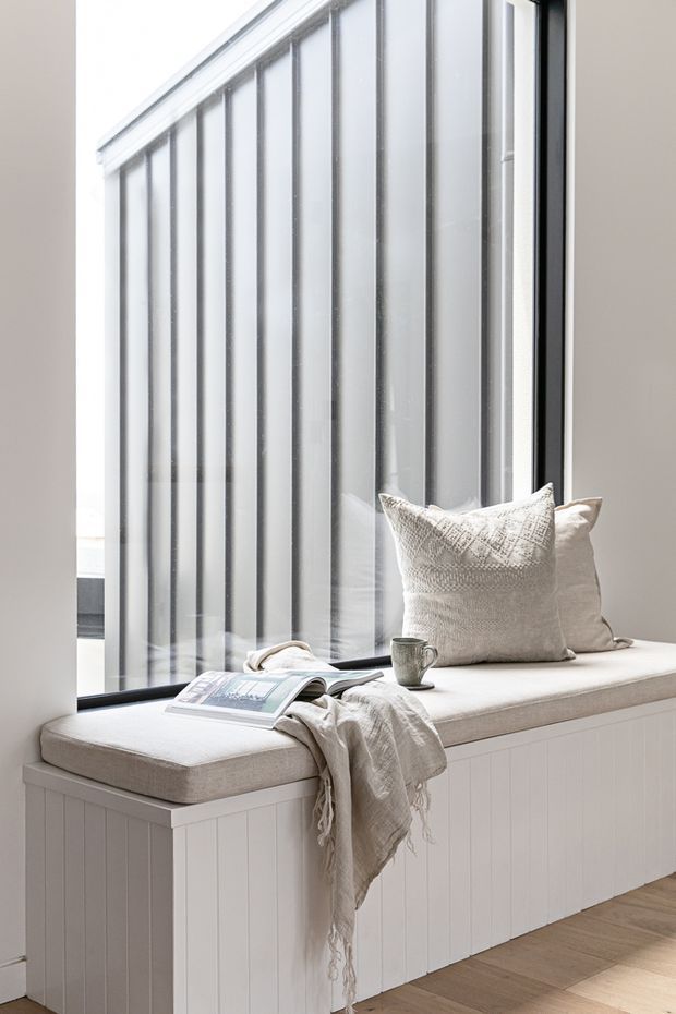 Bright, serene interior detail featuring a built-in window seat beneath tall black-framed glazing. The seat has a white upholstered cushion, textured neutral cushions, a draped throw, and a small cup beside an open magazine. White vertical wall panelling and pale timber flooring create a clean, understated setting.