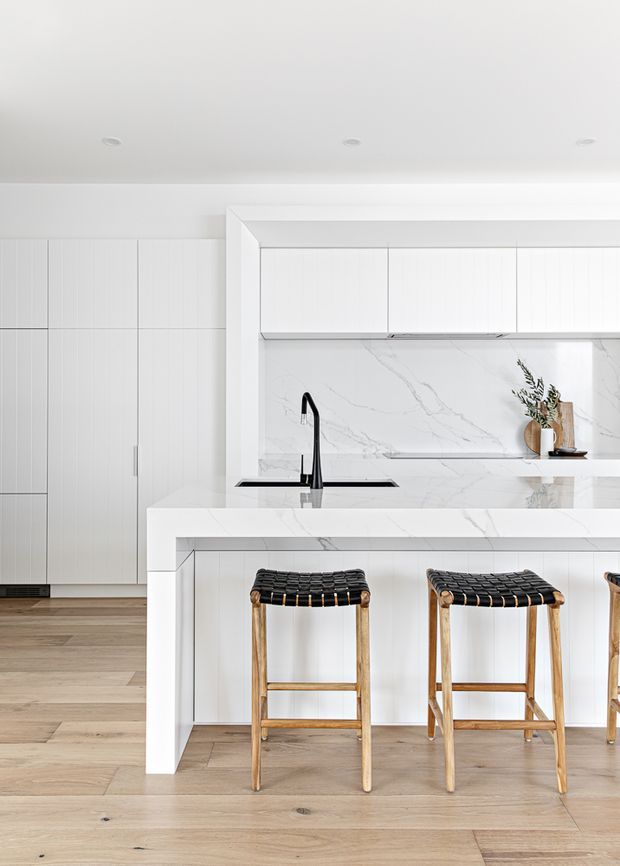 Bright, refined contemporary kitchen with a large white waterfall-edge island, integrated black sink and gooseneck tap, white veined stone splashback and benchtops, and extensive white vertical-panel cabinetry. Timber flooring and woven black-and-timber bar stools add warmth and contrast.