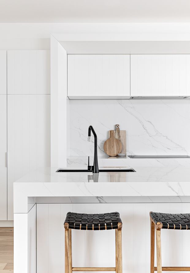 Bright, minimalist white kitchen with a marble-look waterfall island and integrated cooktop, white vertically panelled cabinetry, matching marble-look splashback, and a matte black gooseneck tap. Two woven black-and-timber bar stools sit at the island, adding warm natural contrast.