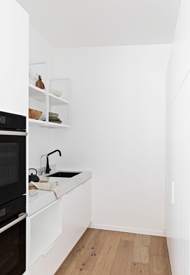 Bright, compact white galley kitchen with integrated black appliances, white flat-panel cabinetry, open shelving, a white stone-look benchtop, black sink and tapware, and warm timber plank flooring. The space is narrow with a clean minimalist fit-out and abundant white surfaces.