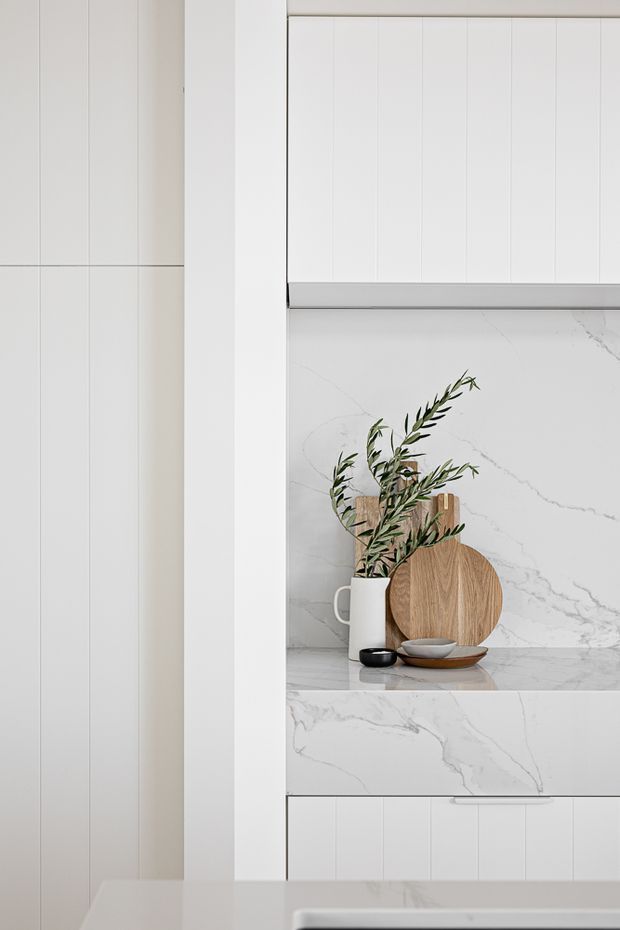 A minimalist white kitchen detail featuring vertically panelled white cabinetry, a white marble-look stone splashback and benchtop with grey veining, and a recessed display niche styled with a white jug, wooden boards, bowls, and olive branches.