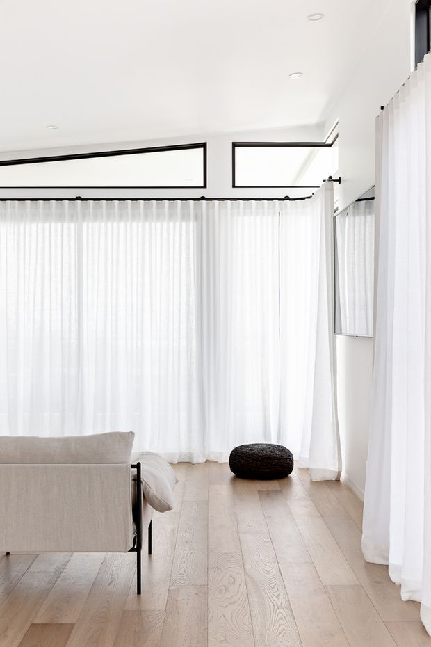 Bright, minimalist living room with pale timber flooring, white sheer floor-to-ceiling curtains, expansive black-framed clerestory and corner glazing, and a light neutral sofa. A low black woven ottoman provides a contrasting accent. The space is airy and softly daylit with clean contemporary detailing.