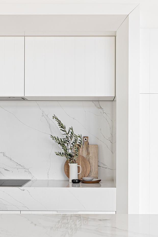 A bright, minimalist white kitchen featuring full-height vertically panelled cabinetry, a white marble-look stone splashback and benchtop with subtle grey veining, and integrated cooking appliances. Timber chopping boards, neutral ceramics, and olive branches provide restrained natural accents.