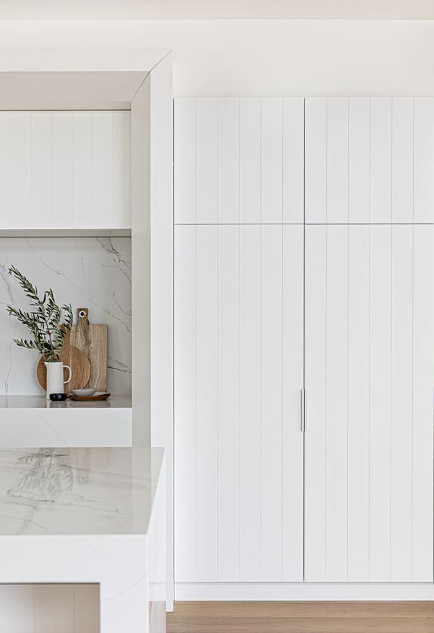 Close-up view of a bright, minimalist white kitchen with tall vertical-panel cabinetry, integrated handles, a white marble-look splashback and benchtop, and an open display niche holding timber boards and a small vase. Light timber flooring is visible at the base.