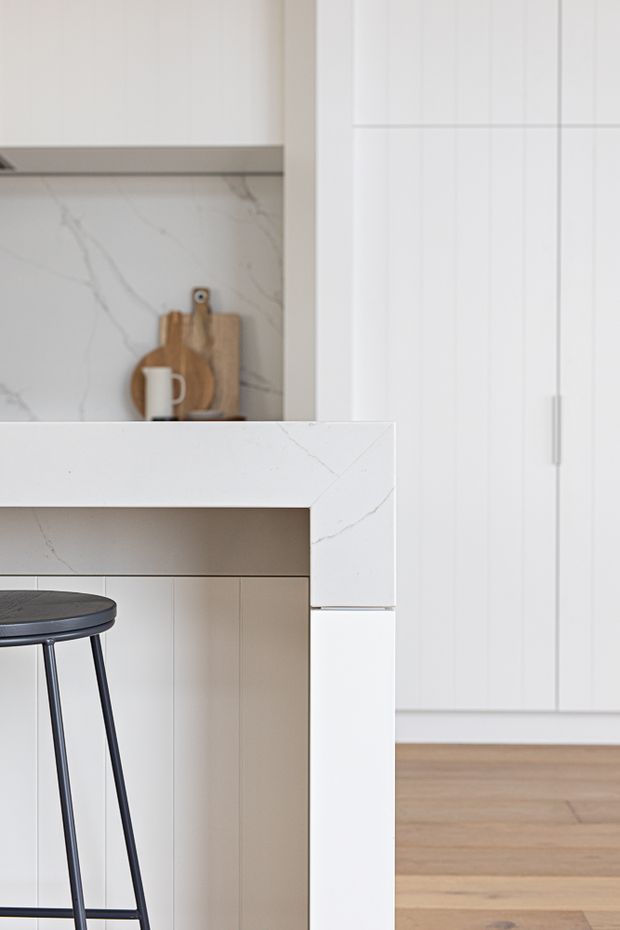Close-up view of a bright, refined kitchen featuring a white veined stone island benchtop with waterfall edge, white paneled cabinetry, a matching stone splashback, light timber flooring, and a dark metal bar stool. Wooden boards and a ceramic jug add subtle styling in the background.