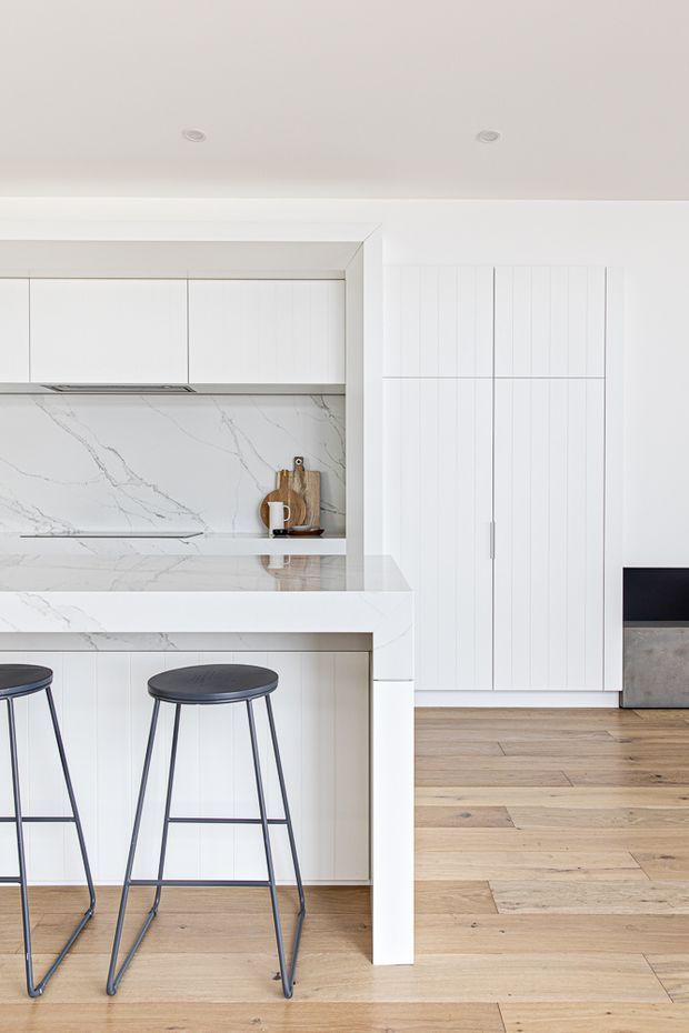 Bright, minimalist white kitchen with a marble-look stone splashback and island benchtop, integrated white cabinetry, vertical panelled cabinet doors, black metal bar stools, and warm timber flooring. The kitchen opens toward an adjacent living area with a low dark-and-grey cabinet visible at right.