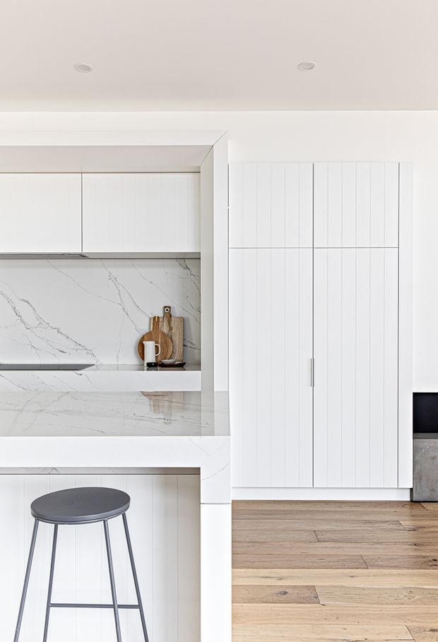Bright, minimalist white kitchen with vertically fluted cabinetry, integrated open shelving, and a marble-look stone splashback and benchtop with prominent veining. A slim dark stool sits at the peninsula, while light timber flooring continues into the adjacent space. The cabinetry and clean-lined built-in joinery create a refined contemporary appearance.