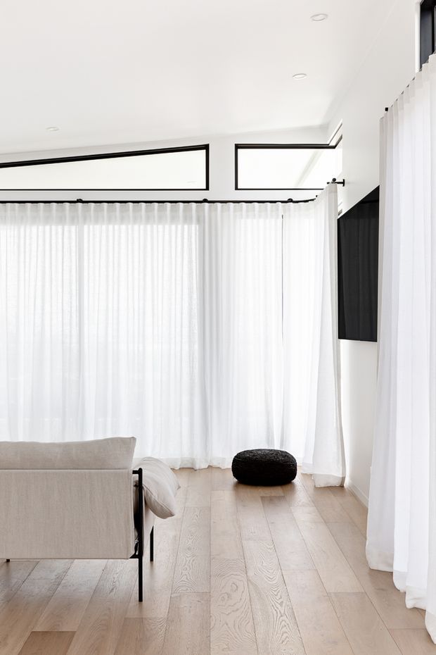 Bright minimalist living room with pale timber flooring, a neutral upholstered sofa, sheer white floor-to-ceiling curtains, high clerestory windows with black frames, recessed ceiling lights, and a small black ottoman. The space has abundant diffused natural light and a clean, restrained palette.