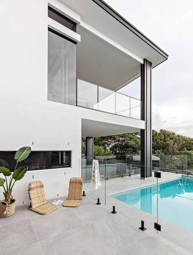 A contemporary multi-level house exterior with a covered balcony and pool terrace. The white rendered facade features large glazing, slim dark metal structural columns, frameless glass balustrades, and a pale grey tiled outdoor deck surrounding a bright turquoise swimming pool. Wicker lounge chairs and tropical planting occupy the foreground.