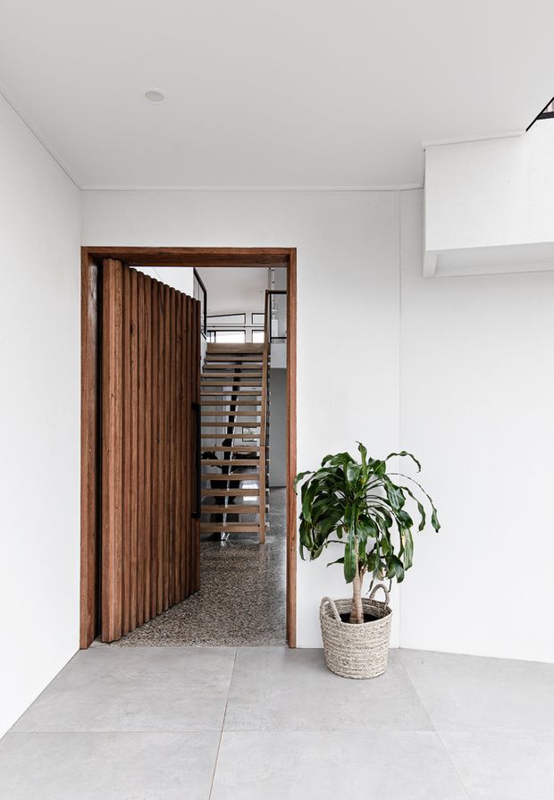Bright minimalist entryway with white walls and ceiling, large pale concrete-look floor tiles, and a dark timber-framed opening. A distinctive vertical timber slat door opens toward a stairway with open-riser timber treads and metal structure beyond. A potted indoor plant sits beside the doorway, and a partial white built-in or projecting architectural element is visible at upper right.