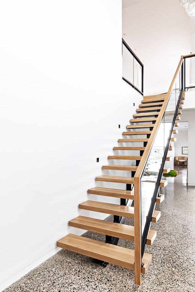 Bright contemporary interior featuring an open-riser staircase with solid timber treads, a timber handrail, and black-framed glass balustrades. The staircase rises beside a plain white wall, with a polished terrazzo floor and glimpses of an open-plan living area beyond.